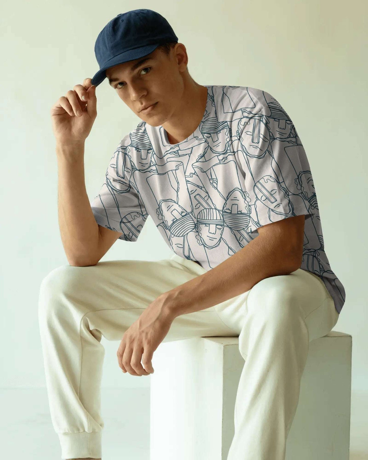 Man wearing a notch patterned shirt and white pants sitting on a white block.